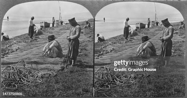 Stereoscopic image showing fishermen mending nets at the entrance of the River Jordan to the Sea of Galilee in Palestine, circa 1900.