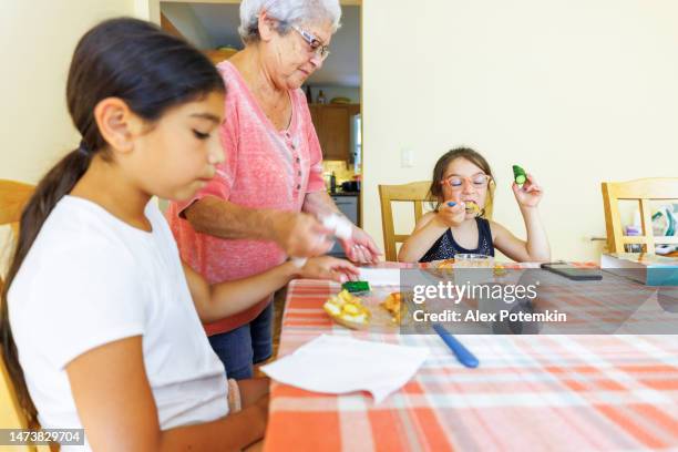 cena in famiglia. nonna che serve cibo ai suoi nipoti. la ragazza più grande è annoiata e di cattivo umore, e la piccola sta mangiando con appetito. - povertà foto e immagini stock