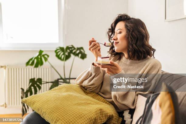 young woman is sitting in the living room alone enjoying taste of the cake - eating cake stock pictures, royalty-free photos & images