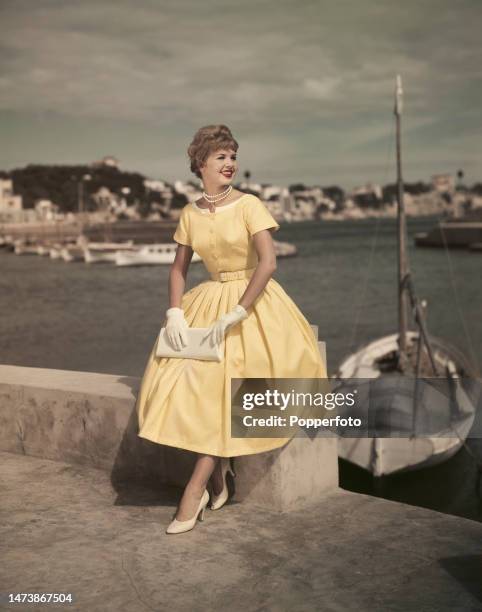 Vacation scene of a female fashion model posed on a harbour wall, she wears a yellow summer frock with a buttoned bodice, full skirt and self fabric...