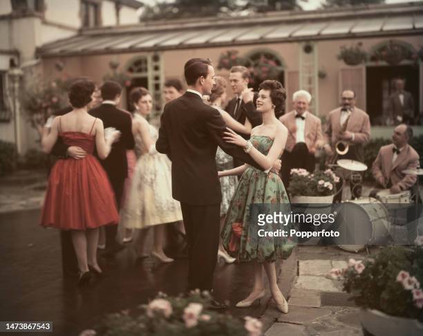 Party scene of young couples dancing to the music of a dance band in the courtyard of a large house, the woman wears a green floral print strapless...