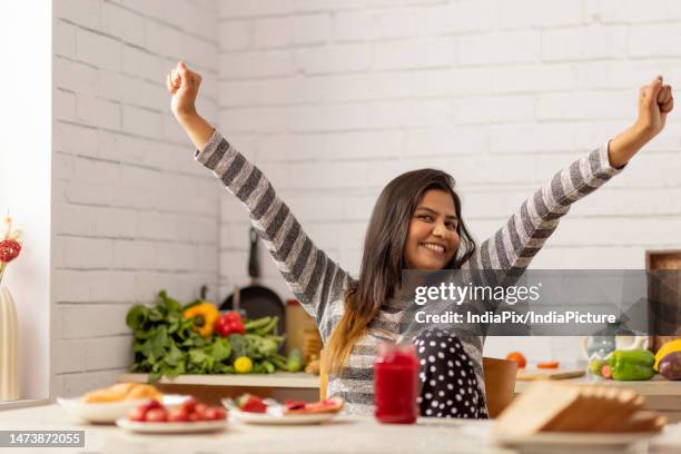 cheerful woman sitting with arms raised in kitchen during breakfast - femme bras tendu cuillère photos et images de collection