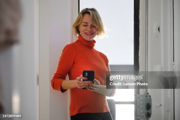 front view shot of a beautiful and confident white woman smiling at the entrance of her house using a smartphone - apartment entry stock pictures, royalty-free photos & images