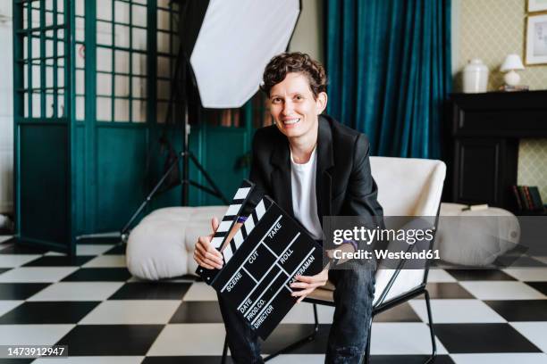 smiling director sitting on chair holding clapboard at film set - studio cinematografico foto e immagini stock