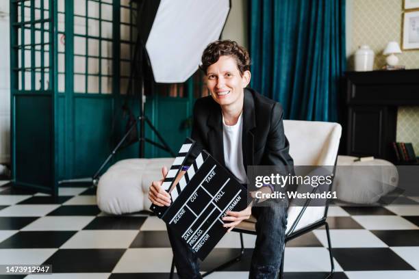 smiling director sitting on chair holding clapboard at film set - filmstudio stockfoto's en -beelden