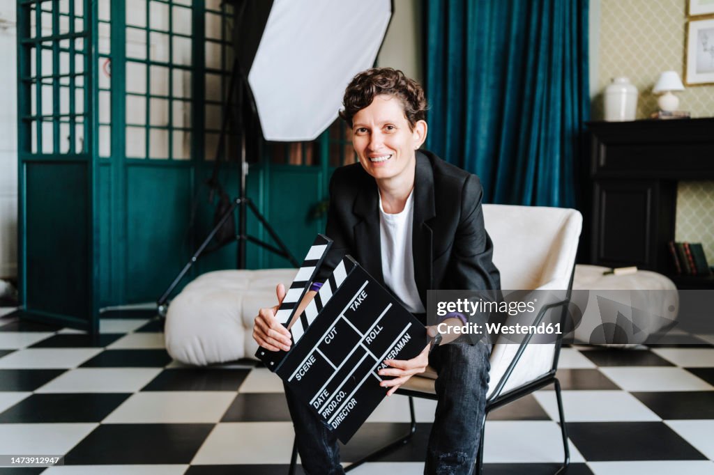 Smiling director sitting on chair holding clapboard at film set