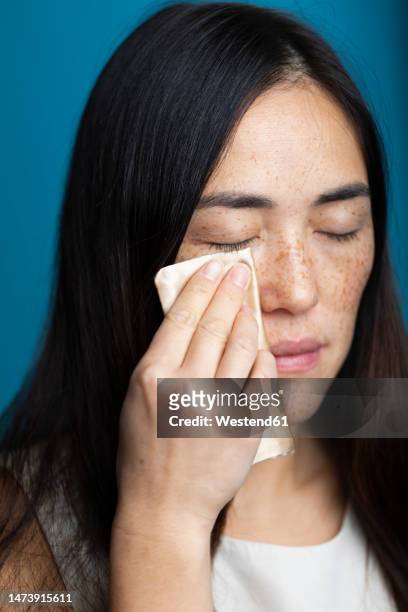 woman with eyes closed cleaning face against blue background - démaquillant photos et images de collection