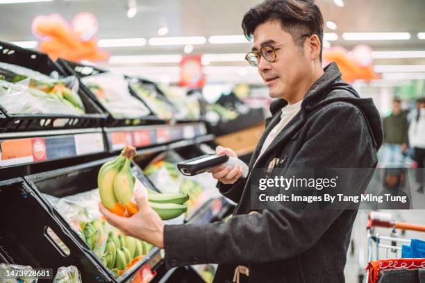 young handsome man using handheld barcode reader scanning a bunch of bananas while shopping in supermarket - supermarket uk stock pictures, royalty-free photos & images