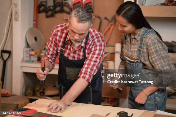 craftsman senior man teaching woman apprentice at woodshop, carpenter training wood for female standing at wood table in workshop, national carpenters day - national teacher stock pictures, royalty-free photos & images