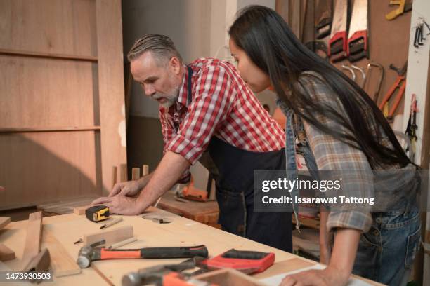 craftsman senior man teaching woman apprentice at woodshop, carpenter training wood for female standing at wood table in workshop, national carpenters day - national teacher stock pictures, royalty-free photos & images