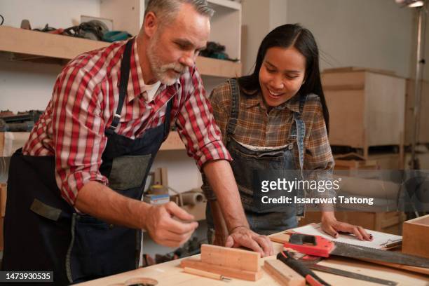 craftsman senior man teaching woman apprentice at woodshop, carpenter training wood for female standing at wood table in workshop, national carpenters day - national teacher stock pictures, royalty-free photos & images