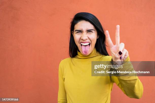 latino woman sticking her tongue out, looking at the camera and giving the peace sign, medium shot - gesto de victoria fotografías e imágenes de stock