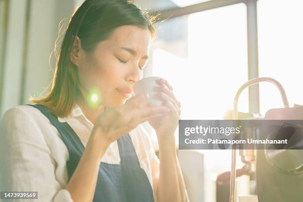 selbstbewusste asiatische japanerin oder barista in der schürze, die an der thekenbar des cafés steht und mit einem lächeln unter sonnenlicht bewaffnet ist und schwarzen kaffee aus weißer tasse riecht. genießen sie das aroma von heißem getränk kaffe - chef smelling food stock-fotos und bilder