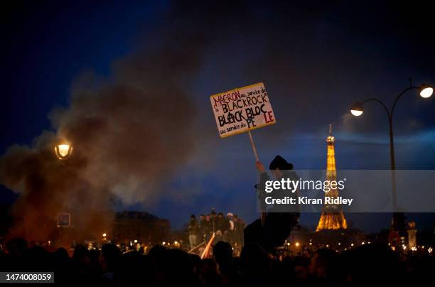 Protester sits on top of a lamp post with a placard that reads "Macron at the service of Black Rock, Black Block at the service of the people" as...