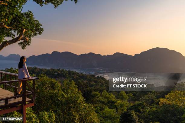 a woman standing at phi phi view point and see the view from a bird's eye view of the mountains, the sea and the sunset. and town on phi phi island, krabi province, thailand - luxury foto e immagini stock