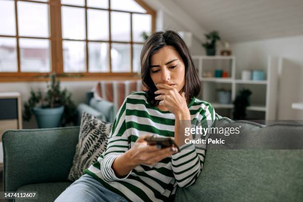 concerned young woman using smart phone in a living room - confused woman stockfoto's en -beelden