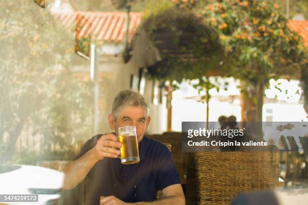 mature man toasting with beer on a bar terrace on vacation - diversión stockfoto's en -beelden