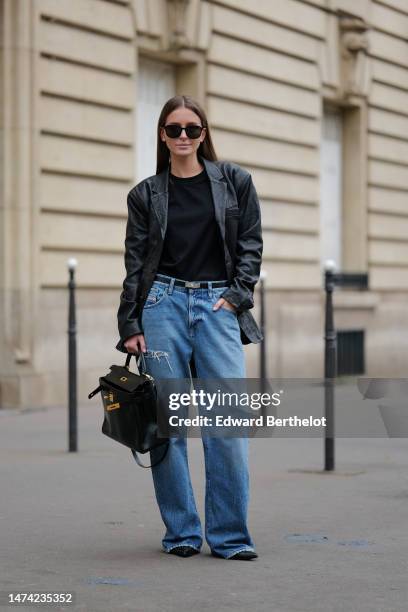 Diane Batoukina wears sunglasses, a black top, a black leather oversized blazer jacket, a Hermes belt, blue denim wide-leg flared jeans / pants, a...