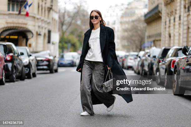 Diane Batoukina wears sunglasses, a black long winter coat, a white t-shirt from Saint Laurent YSL, gray wide-leg flared jeans / denim / pants, a...