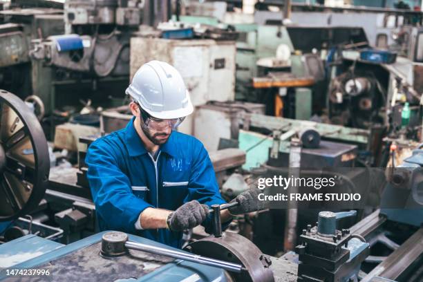 two young workers in blue clothes working on machinery which is a lathe, grinding machine, forming parts and gears in a factory to be assembled into car parts - ingeniería mecánica fotografías e imágenes de stock