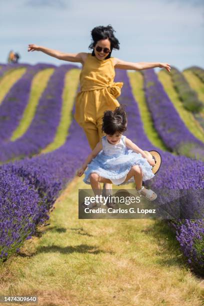 young asian woman wearing a yellow jumpsuit dress and her daughter jump in the middle of a lavender field in  a sunny summer day in kinross, scotland, uk - girls jumpsuit stock pictures, royalty-free photos & images