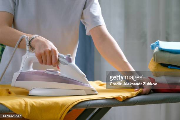 lots of bright, clean, multicolored pressed clothes stacked on the ironing board. a woman or a girl, a housewife, a cleaner or a housekeeper is ironing a t-shirt. the concept of homework, lack of time for household chores. women's duties, home life. - ironing stock pictures, royalty-free photos & images