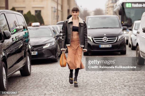 Mary Leest wears gold earrings, a black cropped top, a black shiny leather with beige sheep collar zipper jacket, a camel high waist skirt, black...