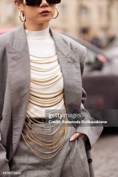 Guest wears black sunglasses, white shoes, white turtle neck top decorated with chains, a grey suit and a beige bag with chains outside the Stella...