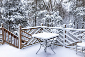 Metal outdoor table and chairs covered in white snow on home house wooden deck with railing fence in northern Virginia winter with forest trees in background