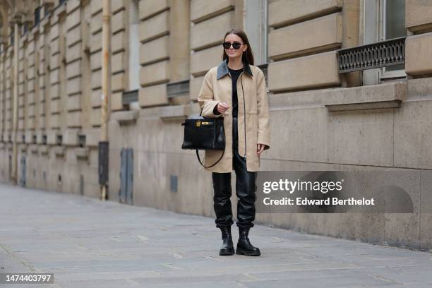 Diane Batoukina wears sunglasses, a beige trench coat with black collar, a black t-shirt, a black leather Hermes bag, black leather shiny pants,...