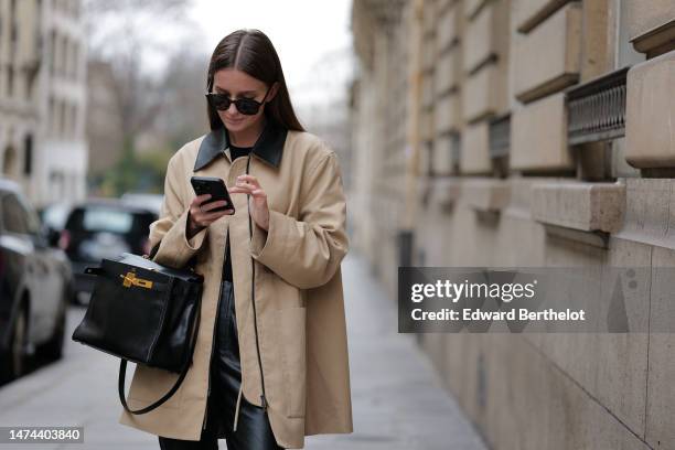 Diane Batoukina wears sunglasses, a beige trench coat with black collar, a black t-shirt, a black leather Hermes bag, black leather shiny pants,...