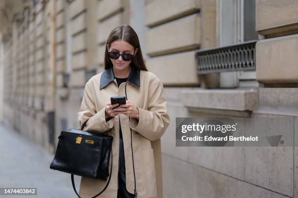 Diane Batoukina wears sunglasses, a beige trench coat with black collar, a black t-shirt, a black leather Hermes bag, black leather shiny pants,...