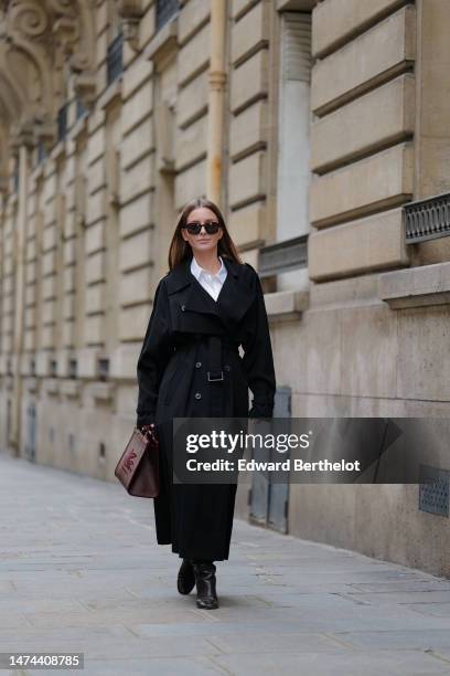 Diane Batoukina wears sunglasses, a white shirt, a black long trench coat, an Etro bag, black leather boots, during a street style fashion photo...