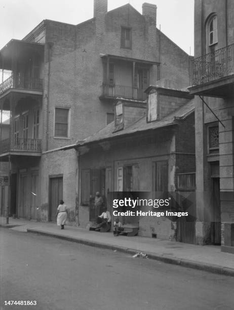 View from across street of a man and child sitting on steps and a woman walking down sidewalk in the French Quarter, New, between 1920 and 1926....