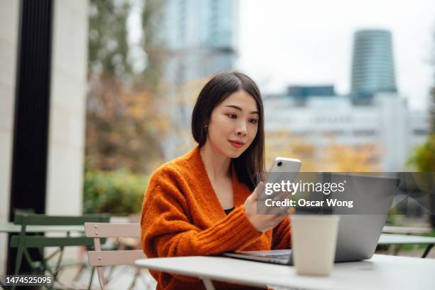 young asian woman talking on mobile phone while working with laptop at sidewalk cafe - flexible working stock pictures, royalty-free photos & images