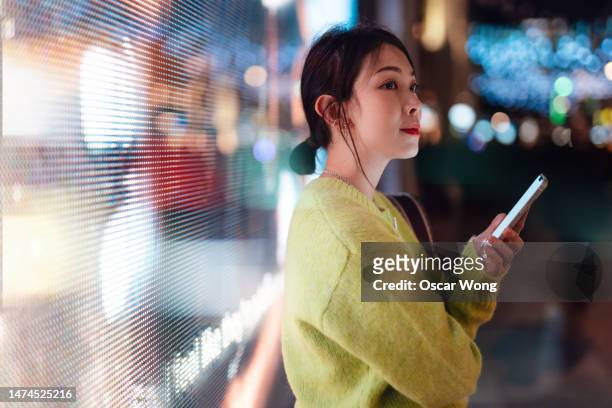 young asian woman using smartphone on city street at night, standing against futuristic illuminated digital display. - mobile billboard stock pictures, royalty-free photos & images