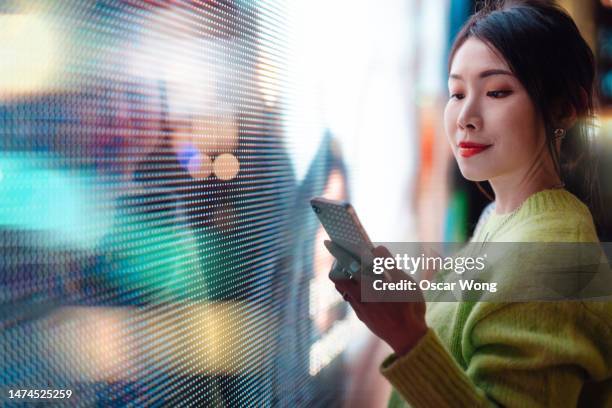 young asian woman using smartphone on city street at night, standing in front of futuristic illuminated digital display. - mobile billboard stock pictures, royalty-free photos & images