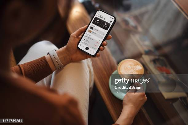 over the shoulder view of young asian woman managing personal banking and finance with online banking app on smartphone, sitting on sofa at home. financial planning. investment. wealth management, banking and finance concept. smart banking with technology - mobiel stockfoto's en -beelden