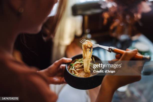 over the shoulder view of young asian woman having lunch in the living room, eating a bowl of noodle soup at home. a hearty and satisfying home-cooked meal. people, food and healthy eating lifestyle - travel stock pictures, royalty-free photos & images