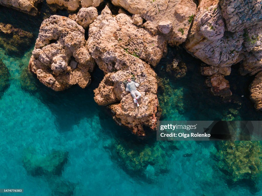 Drone view or selfie of smiling woman with hands behind her head, lying and relaxing on a rock cliff, surrounded by the sea