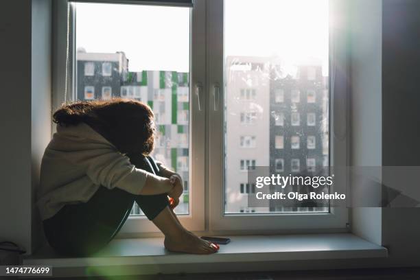 depressed crying woman hiding face on knees, hugging herself on the windowsill against sunny building view. copy space - 頭隠して尻隠さず ストックフォトと画像