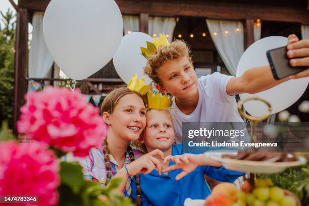 three teen kids in crown caps are making a selfie by phone on birthday party - all age party stock pictures, royalty-free photos & images