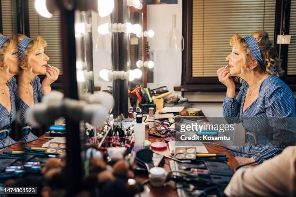a senior actress applies lipstick to her lips while looking in the mirror in a make-up room - theatre dressing room stock pictures, royalty-free photos & images