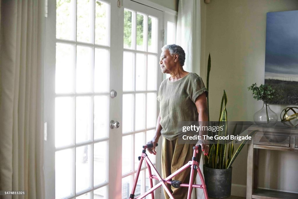 Senior woman looking through window while standing with walker at home