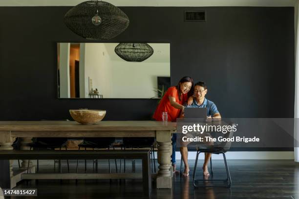 cheerful mature couple looking at laptop in dining room of modern home - mature asian woman candid foto e immagini stock