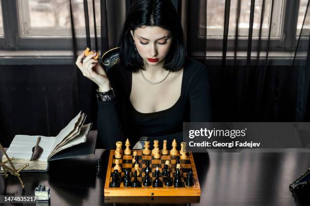 portrait of a beautiful, elegant girl in black, sits behind a chessboard holds a knight piece with a serious, thoughtful expression on her face, staring intently. - chess championship stock pictures, royalty-free photos & images