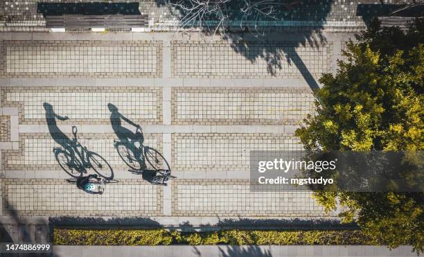 vue aérienne de deux cyclistes et de leurs ombres se reflétant sur la route dans un parc public au coucher du soleil - voie urbaine photos et images de collection