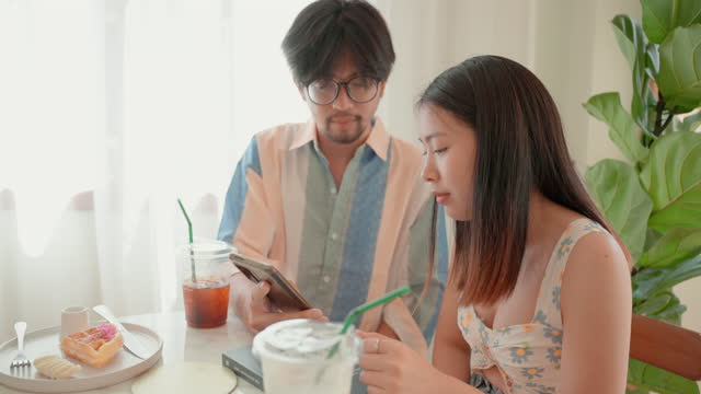 Happy Asian lover having a break in cafe. Young loving couple sitting in a cafe drinking coffee and enjoying surfing internet together. Young Asian couple discussing posts or photos on social media when enjoying romantic date in coffee restaurant.