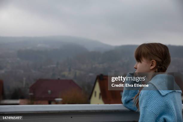 fed up child leaning on elbow against a balcony barrier on a dull, overcast day and looking out across a residential area of a small town surrounded by hills and patches of forest. - leaning on elbows stock pictures, royalty-free photos & images