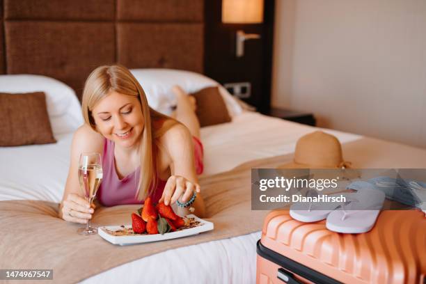 joven y hermosa turista rubia acostada en la cama del hotel bebiendo el champán de bienvenida y comiendo deliciosamente fresas frescas después de llegar a la suite del hotel - equipaje de mano fotografías e imágenes de stock